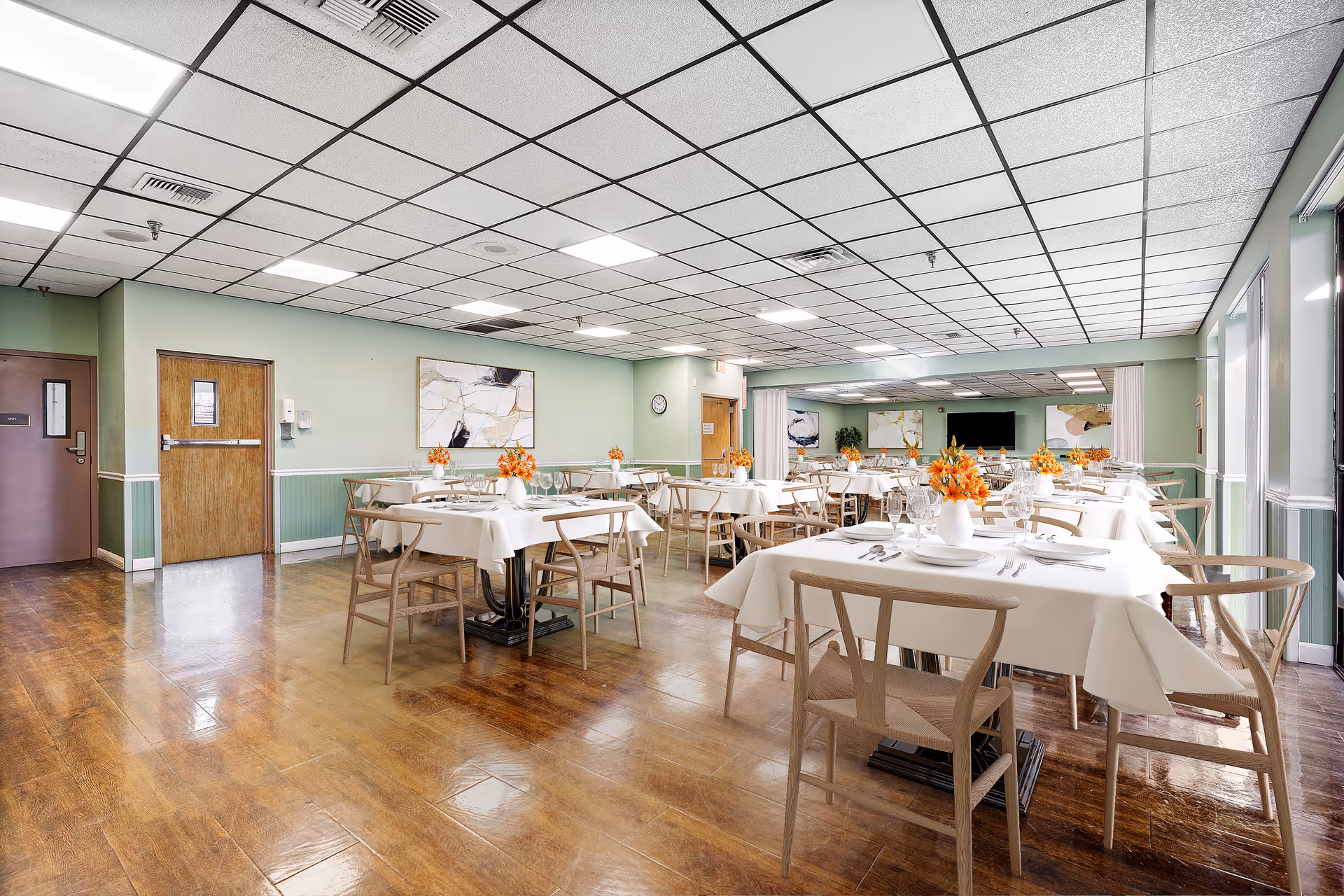 A bright and spacious dining room with several tables covered in white tablecloths, each set with plates, glasses, and silverware. Each table has a vase with orange flowers. The room has light green walls, wooden flooring, and a ceiling with fluorescent lights. There are wooden chairs around the tables and abstract artwork on the walls.