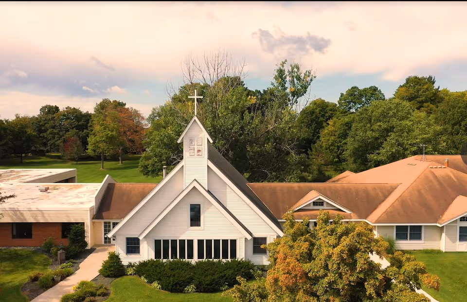 Aerial view of a white chapel with a cross on top, surrounded by green trees and a building with a brown roof, under a partly cloudy sky.