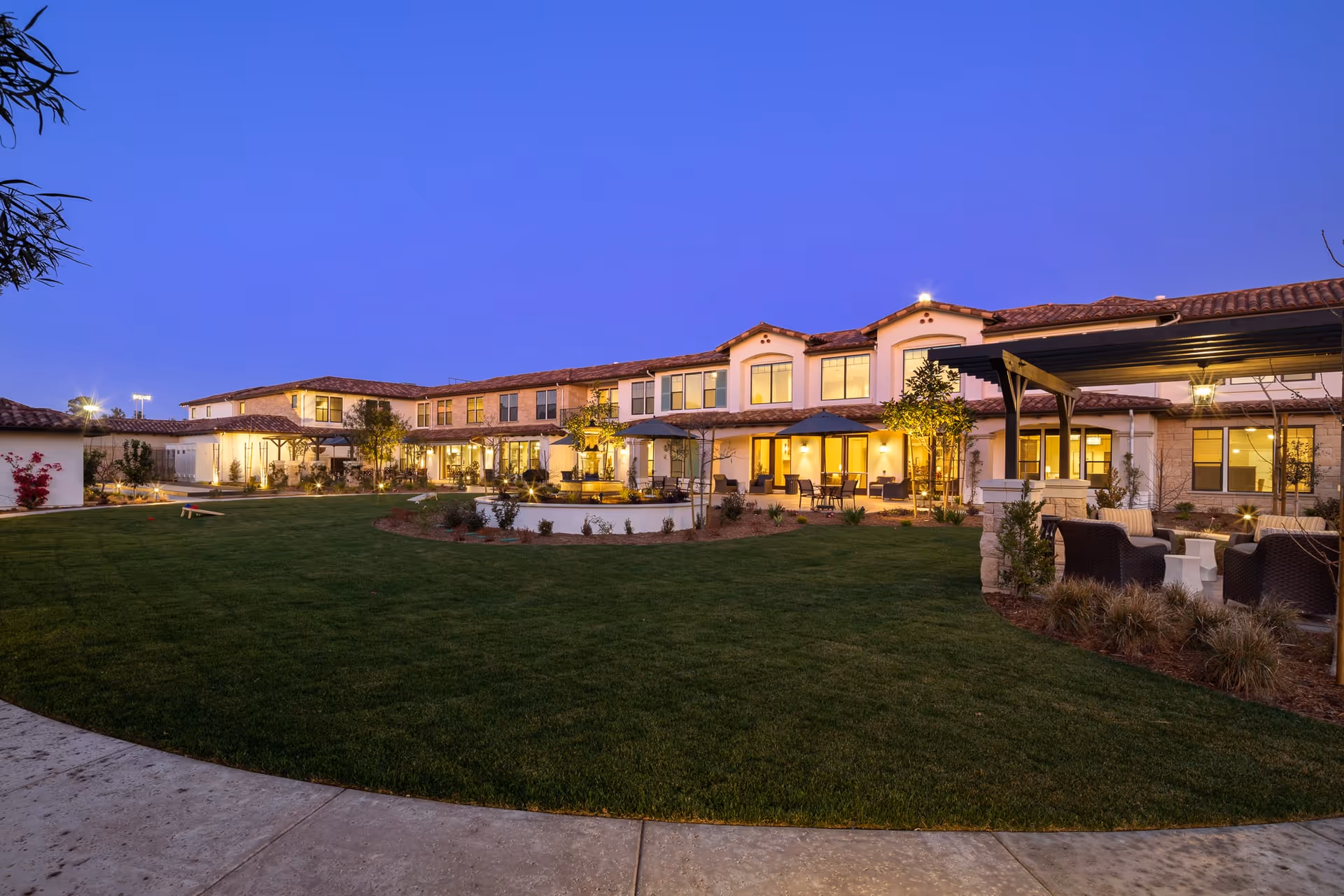 Exterior view of Oakmont of Camarillo senior living facility at dusk, showing a two-story building with warm interior lights, a well-maintained lawn, outdoor seating areas with umbrellas, and a covered patio with chairs and a table.