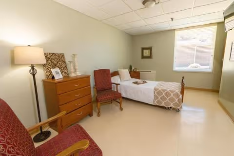 A simple, clean bedroom in a healthcare facility featuring a single bed with white and beige bedding, a wooden dresser with decorative items, two red upholstered chairs, a floor lamp, and a window with blinds letting in natural light.