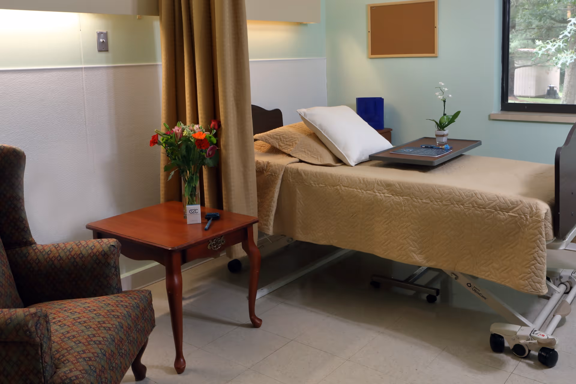 A hospital-style bedroom with a single bed covered in a beige quilt, a white pillow, and a tray table with a small potted plant. Next to the bed is a wooden side table with a vase of colorful flowers and a remote control. There is a patterned armchair beside the table, a window showing greenery outside, and a beige curtain partially drawn.