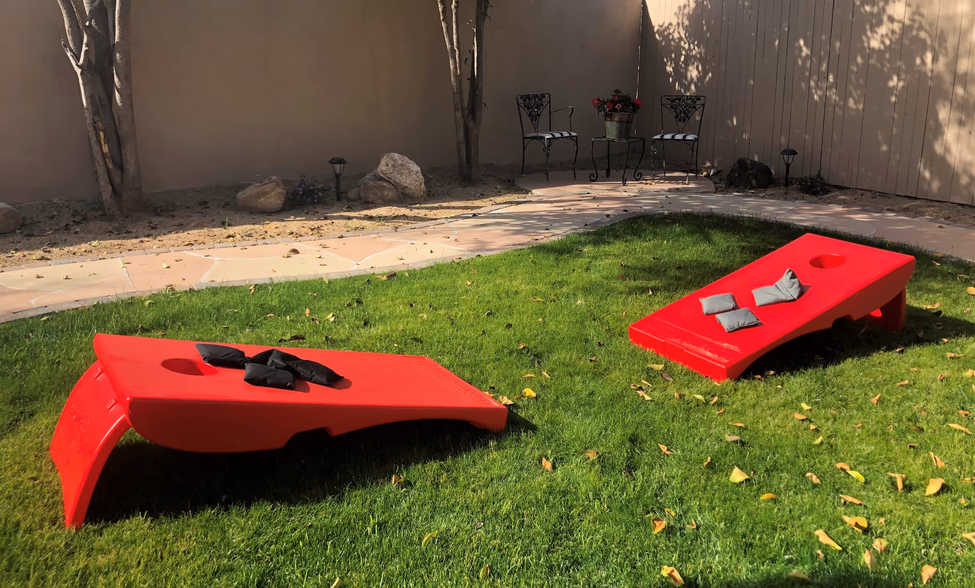 Outdoor grassy area with two red cornhole boards set up for a game, each with bean bags on top. In the background, there is a stone pathway leading to a small seating area with two wrought iron chairs and a small table holding a potted plant. The area is enclosed by a beige wall and wooden fence, with some trees and rocks along the edge.