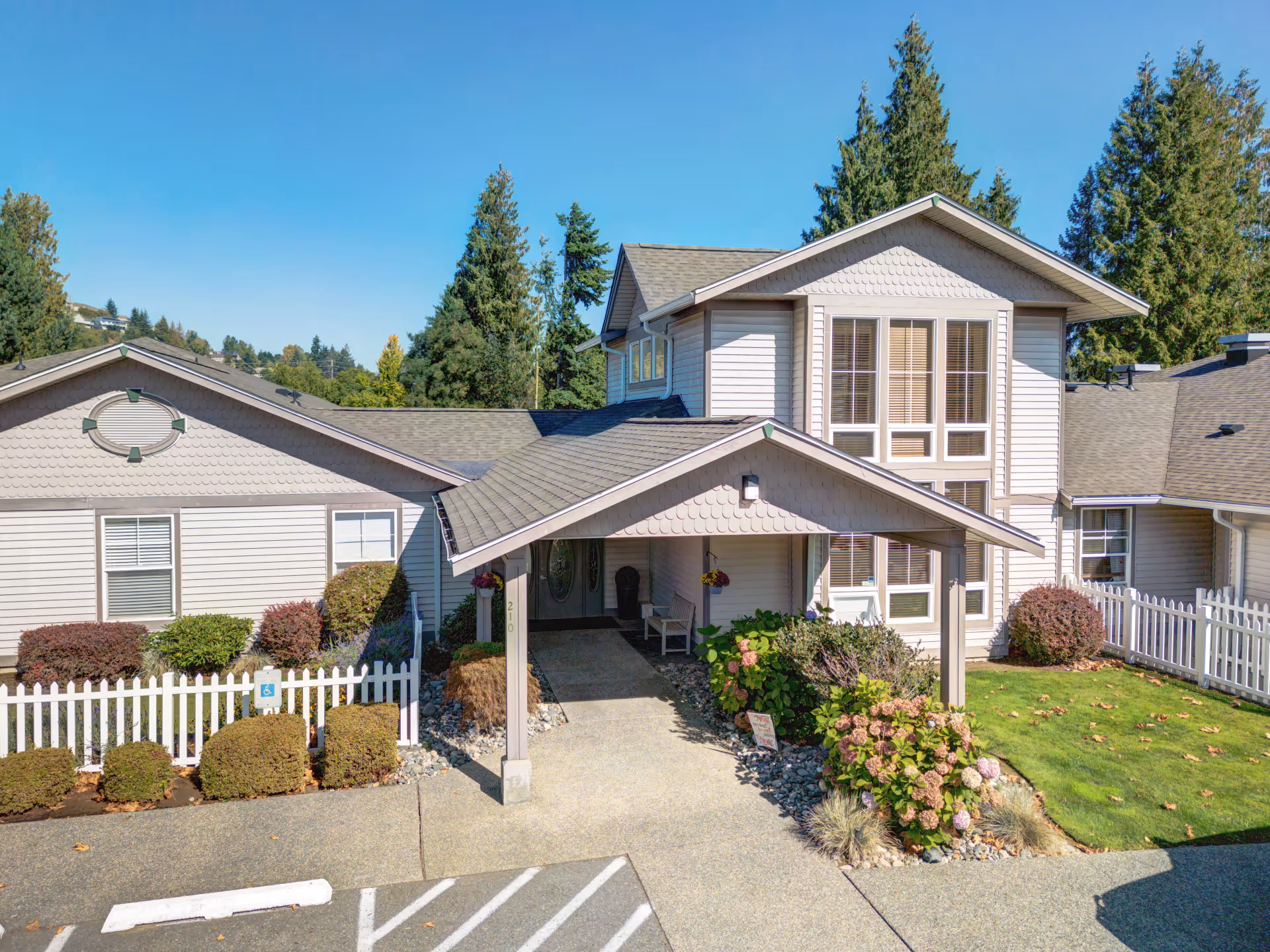 Exterior view of a senior living facility building with beige siding, a covered entrance, white picket fences, landscaped bushes, and a clear blue sky with tall evergreen trees in the background.