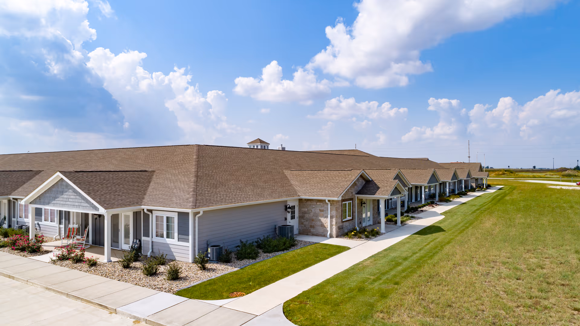 Long single-story assisted living building with multiple covered entrances, a sidewalk and manicured lawn under a partly cloudy sky.