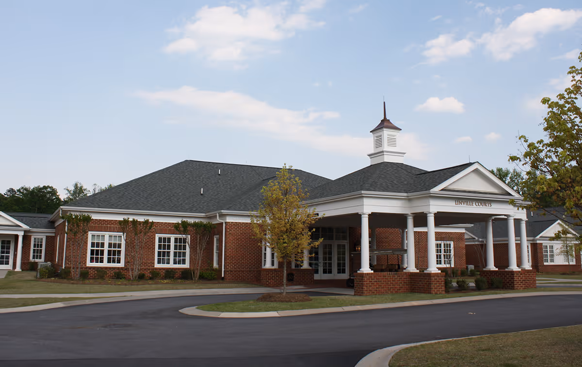Exterior view of a single-story brick building with white columns supporting a covered entrance. The building has multiple windows and a small cupola on the roof. There is a driveway and some landscaping with small trees and bushes around the building.