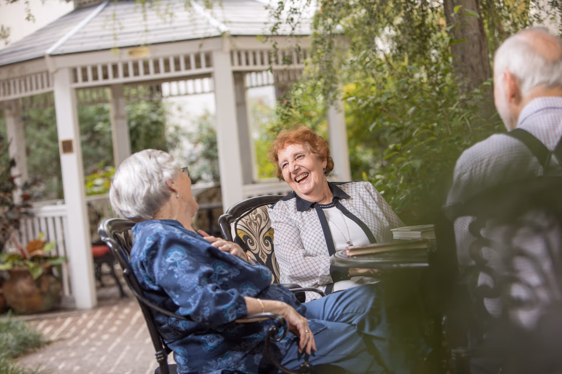 Three elderly people sitting and chatting happily at an outdoor table in a garden area with greenery and a white gazebo in the background.