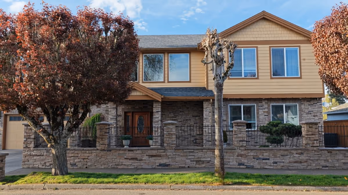 Two-story tan and stone senior living facility with trees and a gated front yard.