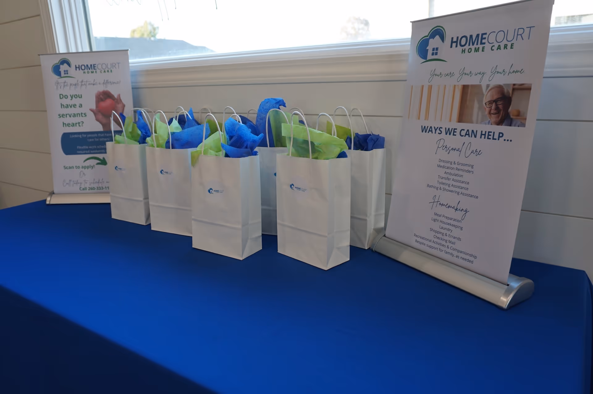 Row of white gift bags with blue tissue on a blue table beside HomeCourt banners.