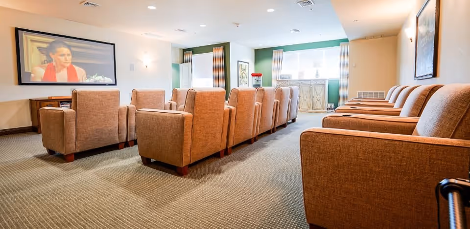 A bright common media room with rows of upholstered armchairs facing a wall-mounted large screen.
