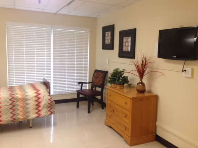 A tidy bedroom in a senior facility featuring a bed, dresser, chair, and mounted TV.