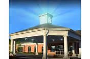 Entrance of a senior living facility with a covered drop-off area supported by white columns, a brick exterior, and a cupola on the roof under a clear blue sky.