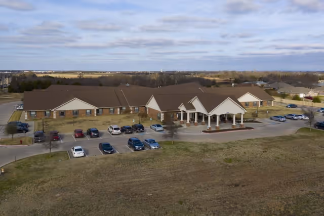 Aerial view of a single-story senior healthcare building with a covered main entrance, parked cars, and surrounding grassy grounds.