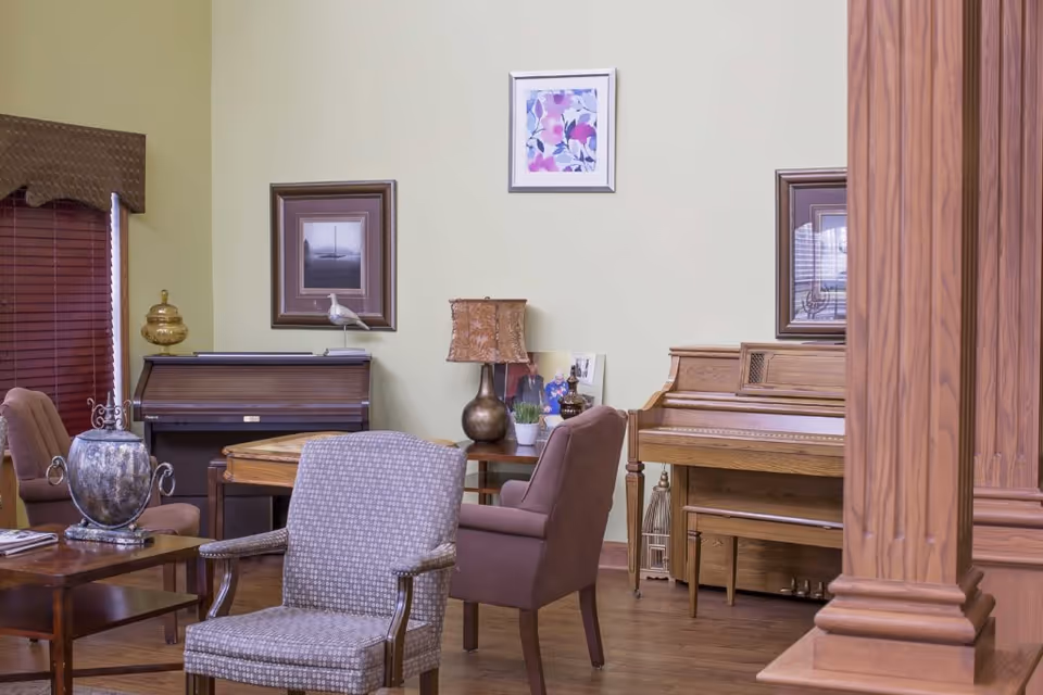 A cozy senior living lounge with chairs, a piano, lamps, and framed artwork.