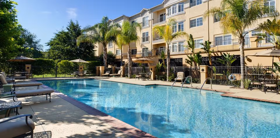Outdoor swimming pool area at a senior living facility with lounge chairs, tables with umbrellas, palm trees, and a multi-story beige building in the background under a clear blue sky.