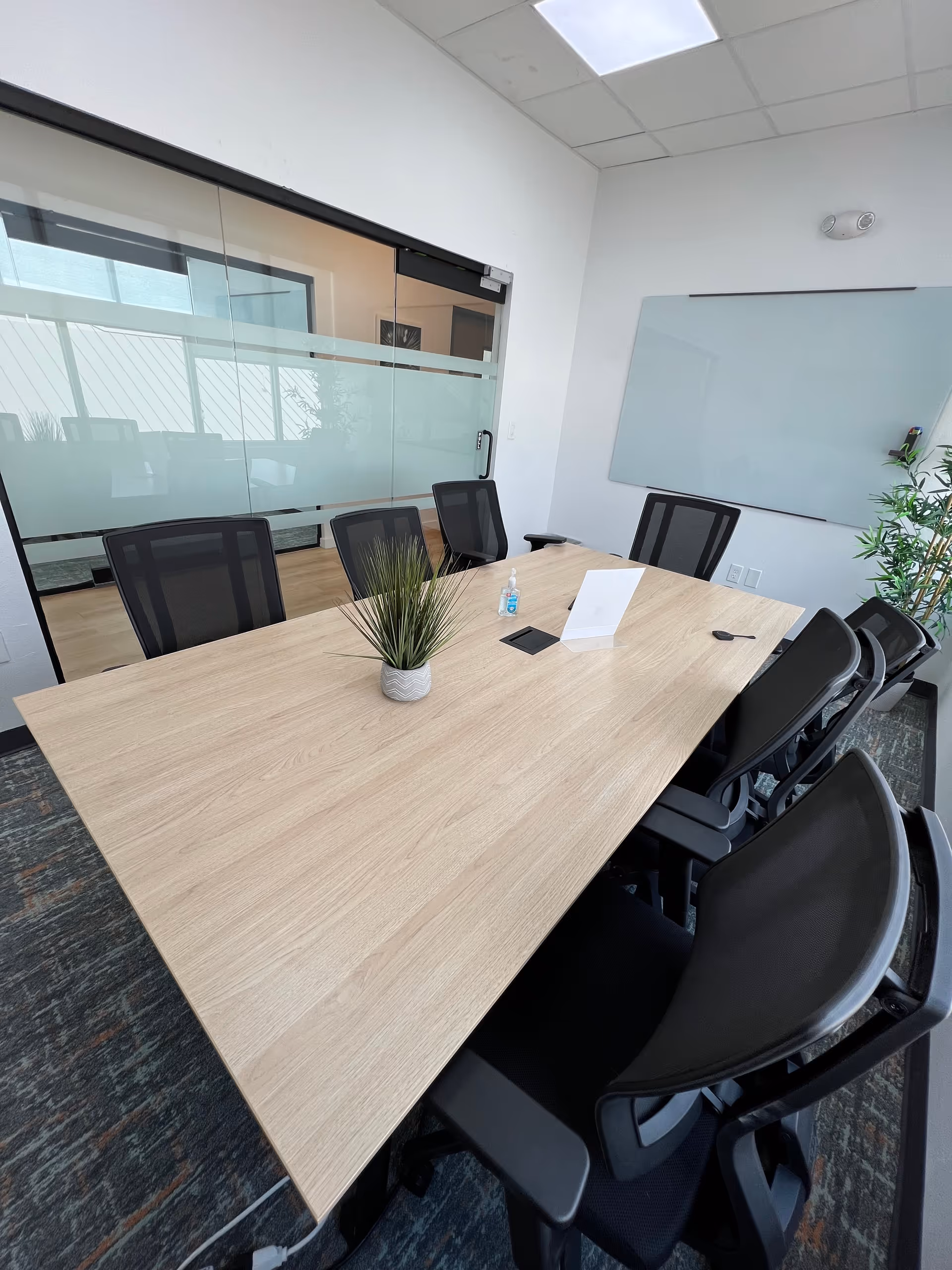 A modern conference room features a large wooden table, black chairs, glass partition, and a whiteboard.