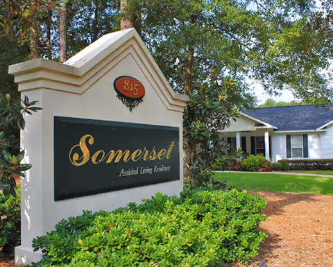 Outdoor view of the entrance sign for Somerset Assisted Living Residence surrounded by green bushes and trees, with a white building and well-maintained lawn in the background.