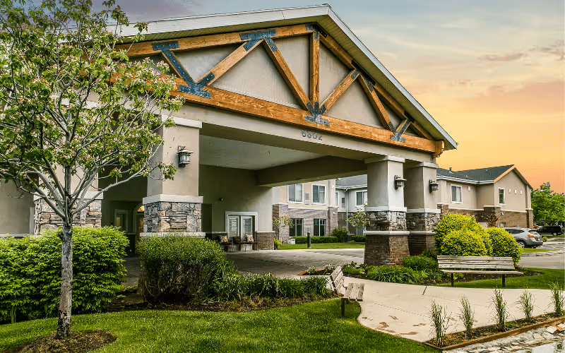Exterior view of Legacy House of Taylorsville showing the entrance with a covered driveway supported by stone and wooden beams. There are green bushes, a tree, and benches along a paved walkway in the landscaped front area. The building is two stories with beige walls and a sunset sky in the background.