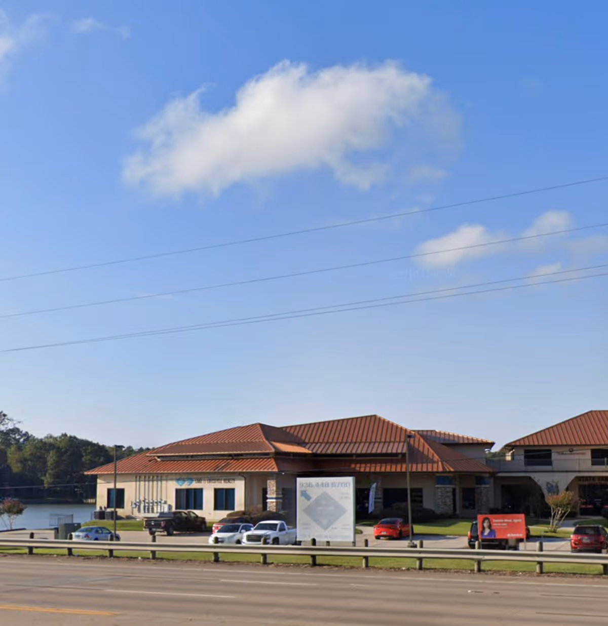 Exterior view of a single-story facility with a red tile roof, parked cars, and road.