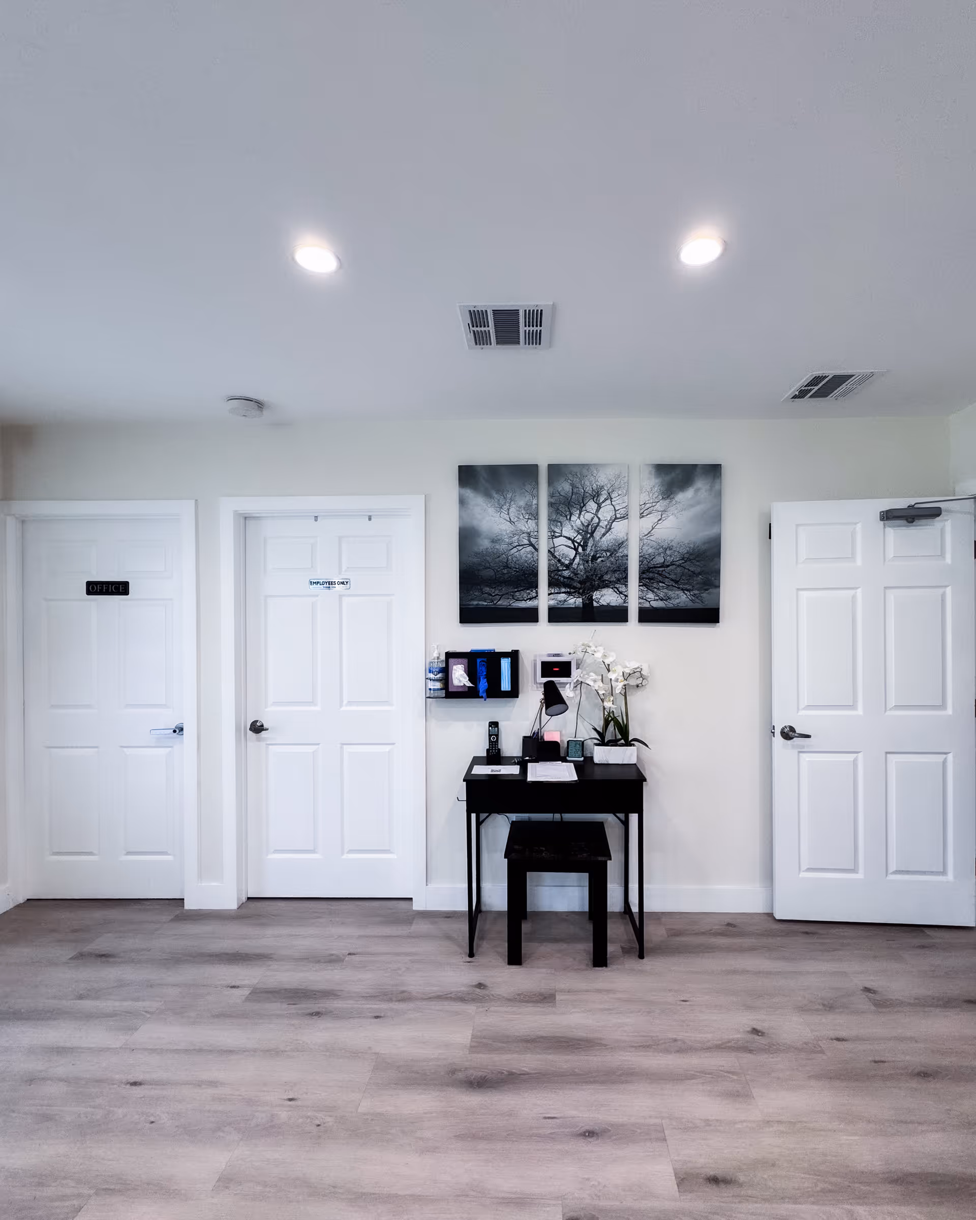 Interior view of a hallway in an assisted living facility with three white doors labeled 'Office' and 'Employees Only'. A small black desk with a stool is placed against the wall, decorated with a white orchid plant, a desk lamp, and some office supplies. Above the desk is a three-panel black and white artwork of a tree. The floor is light wood, and the ceiling has recessed lighting and air vents.
