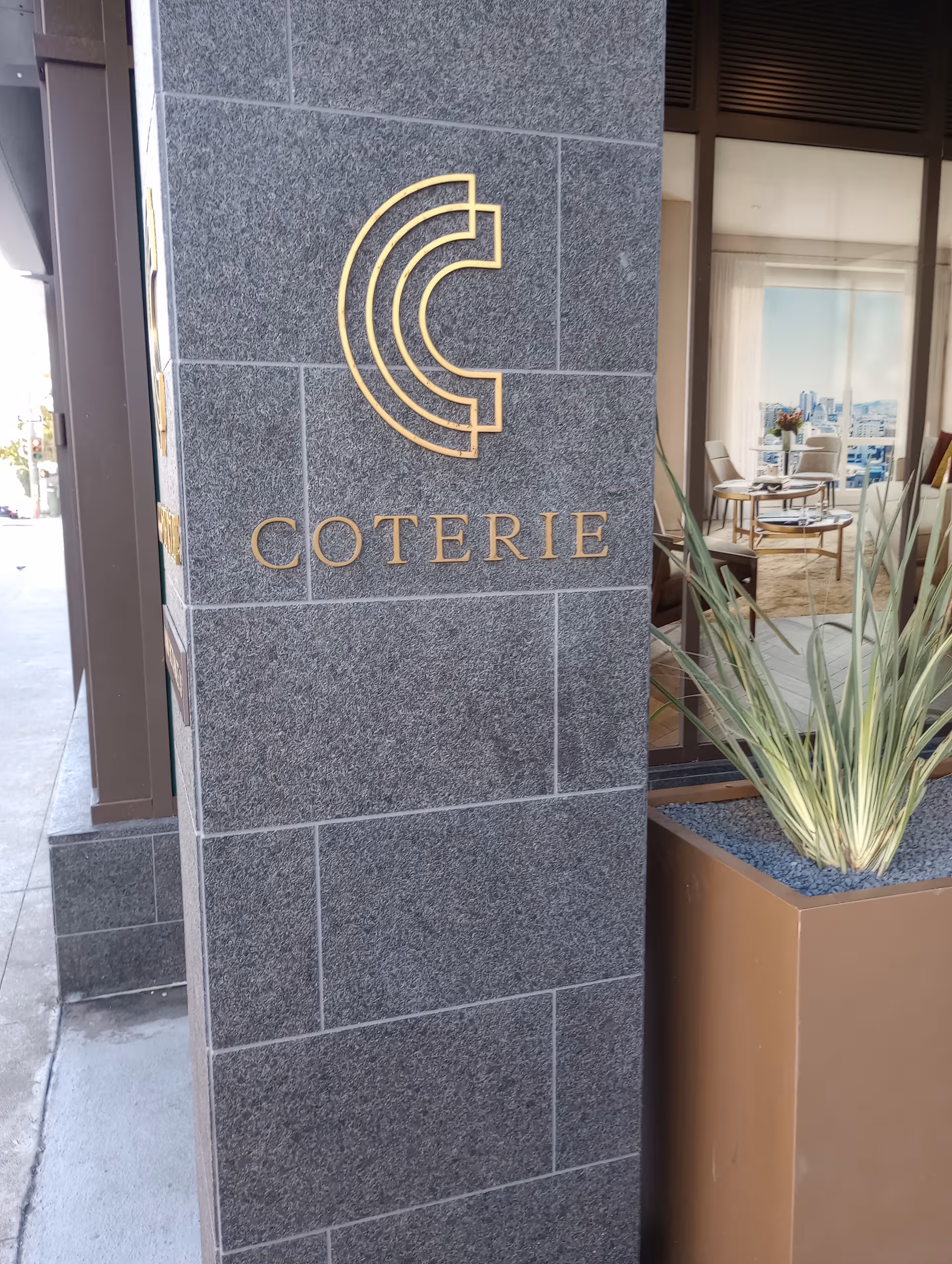 Stone building column with gold Coterie logo beside glass entrance doors.