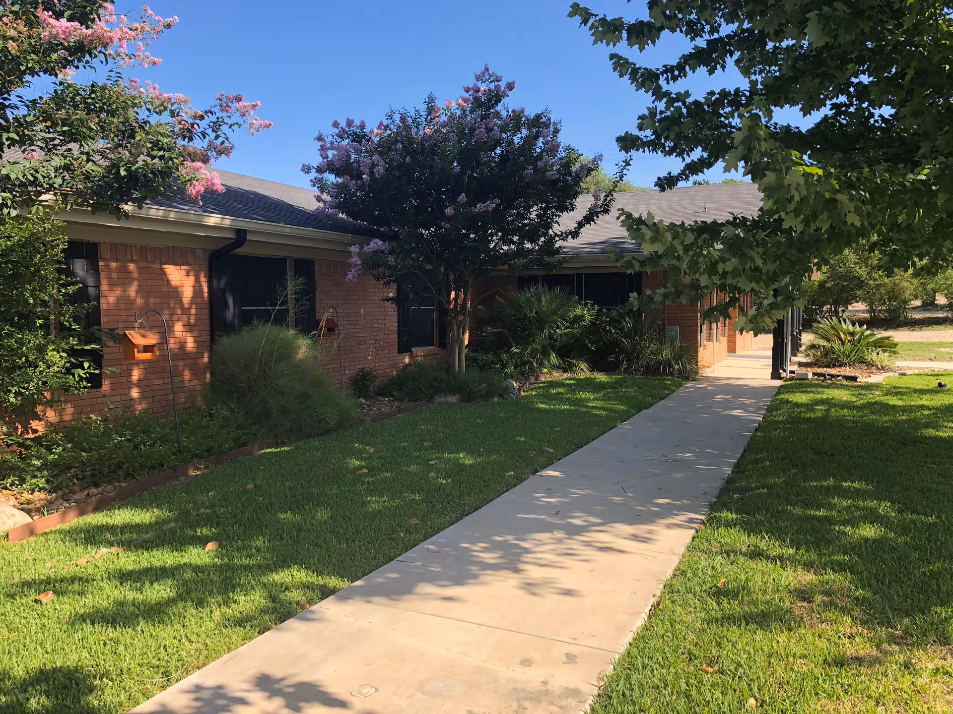 A single-story brick building with a gray roof surrounded by green grass, trees, and flowering bushes under a clear blue sky. A concrete walkway leads to the entrance of the building.