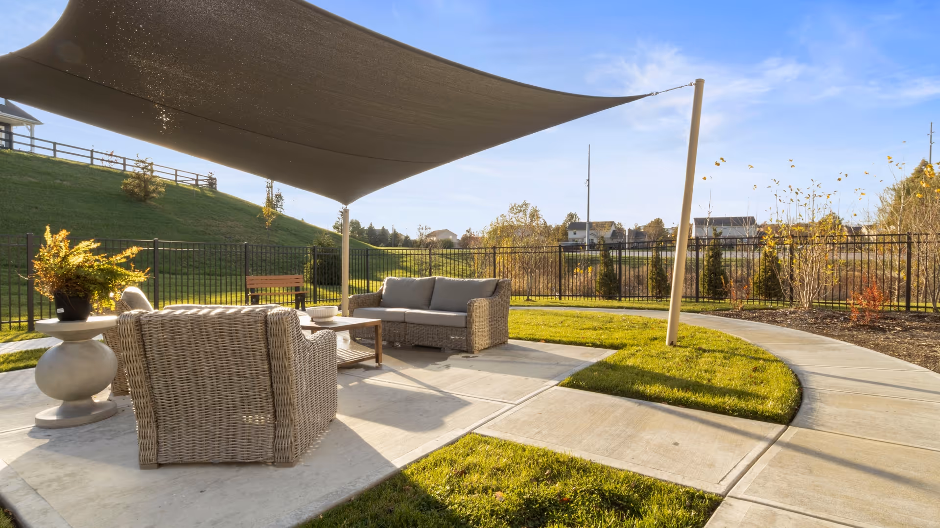 Outdoor seating area with wicker chairs and a sofa under a large shade sail, surrounded by a curved concrete walkway and green grass, with a black metal fence and houses in the background under a clear blue sky.