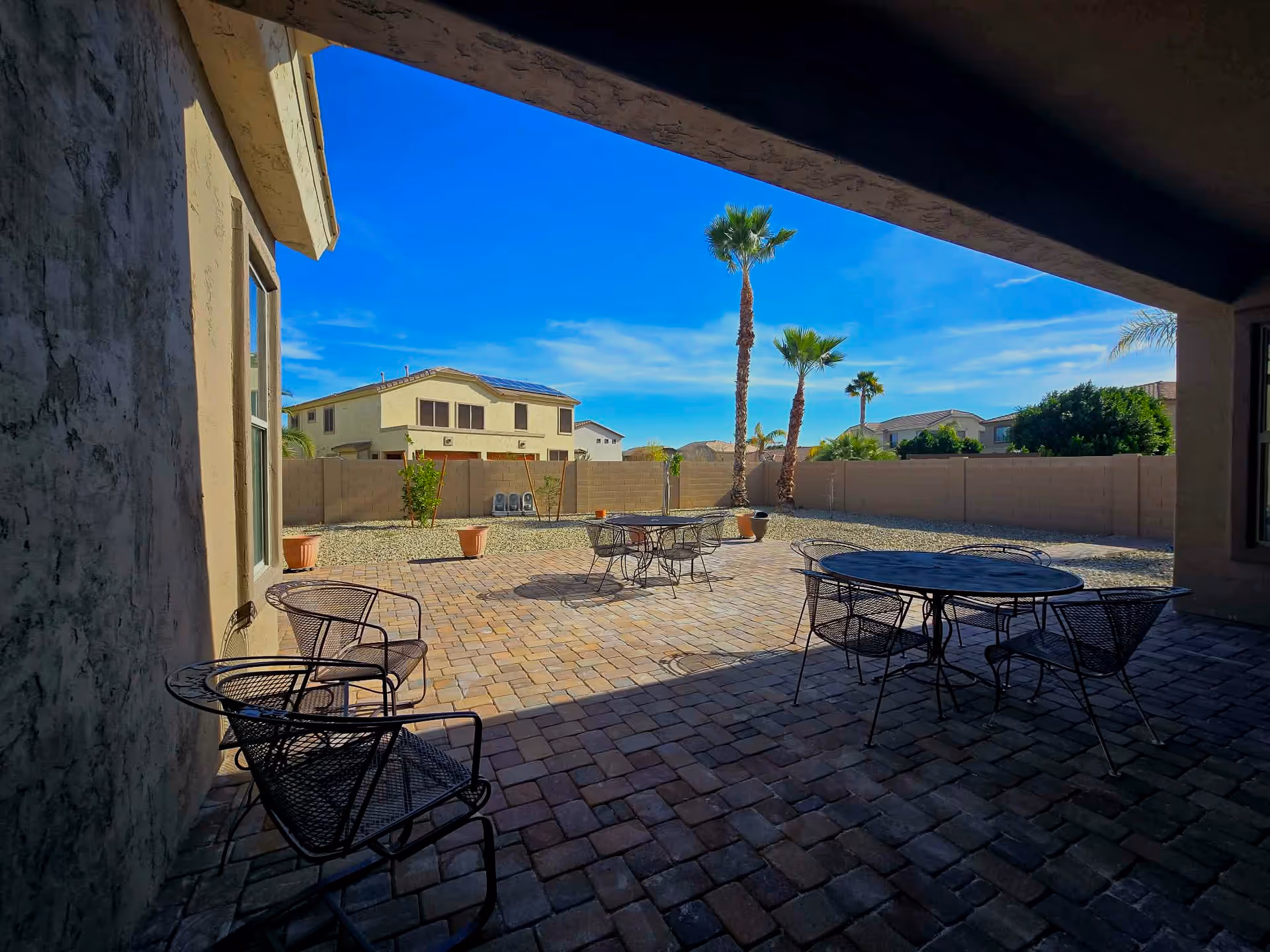 Sunlit paved courtyard with metal tables and chairs, palm trees, and surrounding beige privacy fence.
