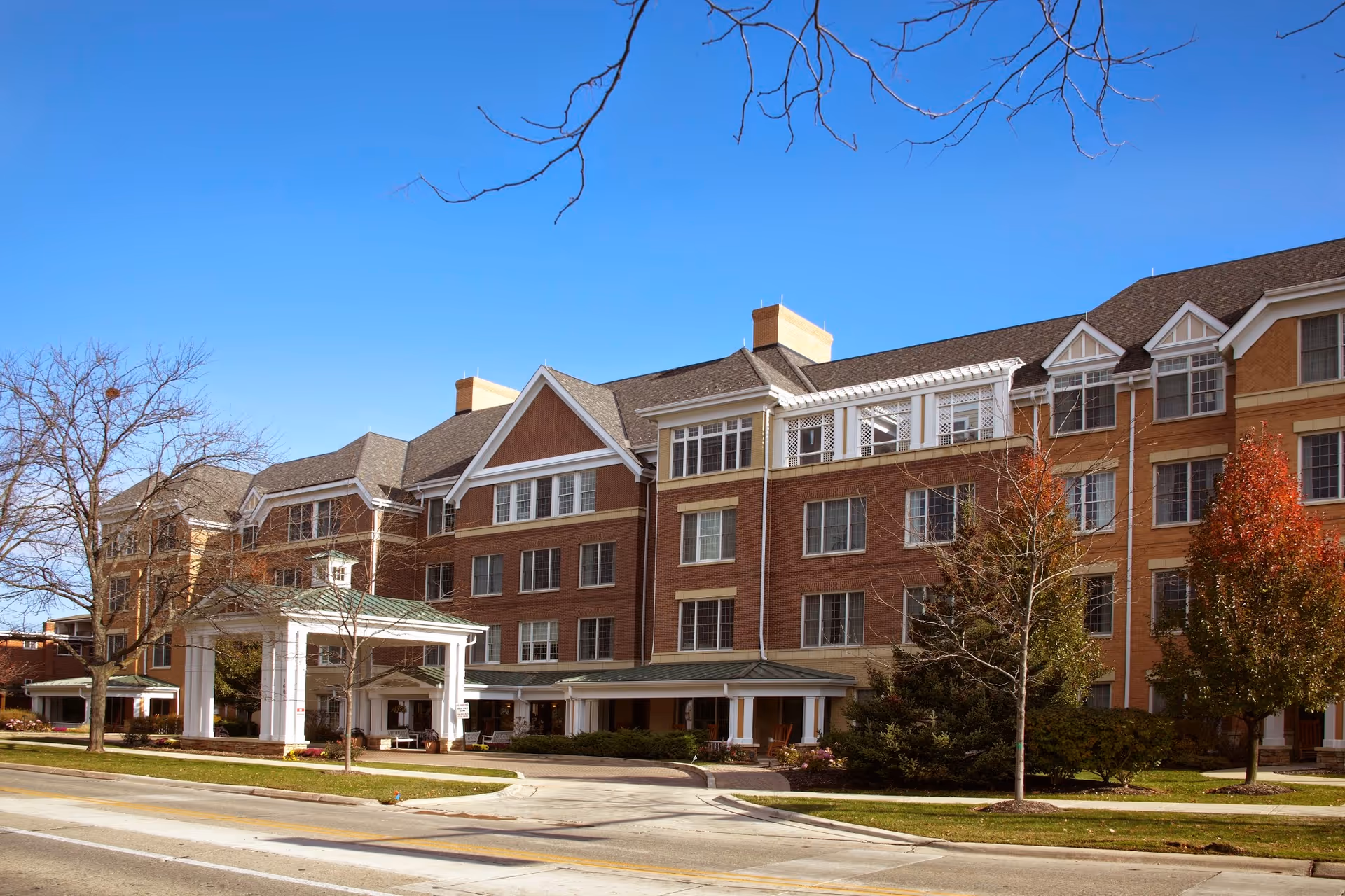 Exterior view of a multi-story senior living facility building with brick and beige siding, large windows, and a covered entrance. Trees with autumn foliage and a clear blue sky are visible.
