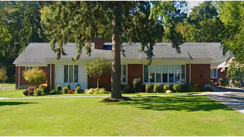 Single-story brick and siding building with a gray roof surrounded by green grass, trees, and shrubs under a clear sky.