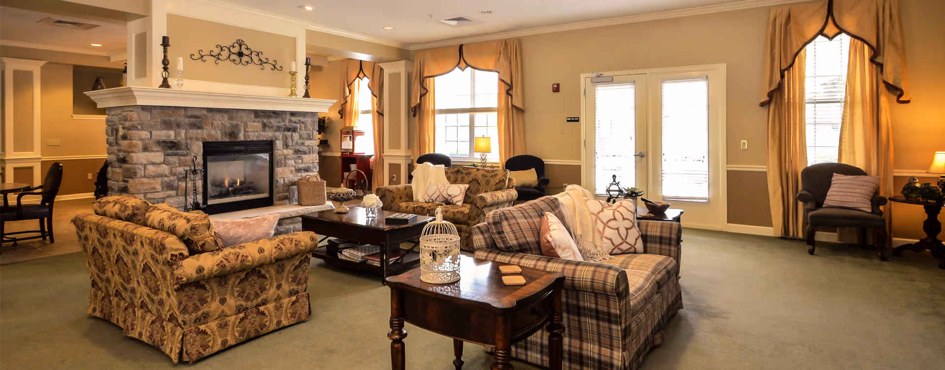 A cozy living room area in a senior living facility with patterned sofas, armchairs, a stone fireplace, and large windows with yellow curtains allowing natural light to fill the room. There are side tables with decorative items and lamps providing warm lighting.