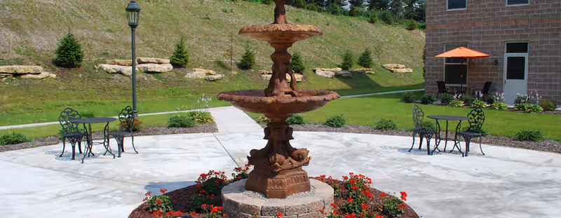 Outdoor patio area with a large decorative two-tiered fountain surrounded by red flowers in the center. There are black metal tables and chairs on the concrete patio, a lamppost, and a building with an orange umbrella over a seating area in the background. The area is landscaped with grass, small shrubs, and a hillside with rocks and trees.