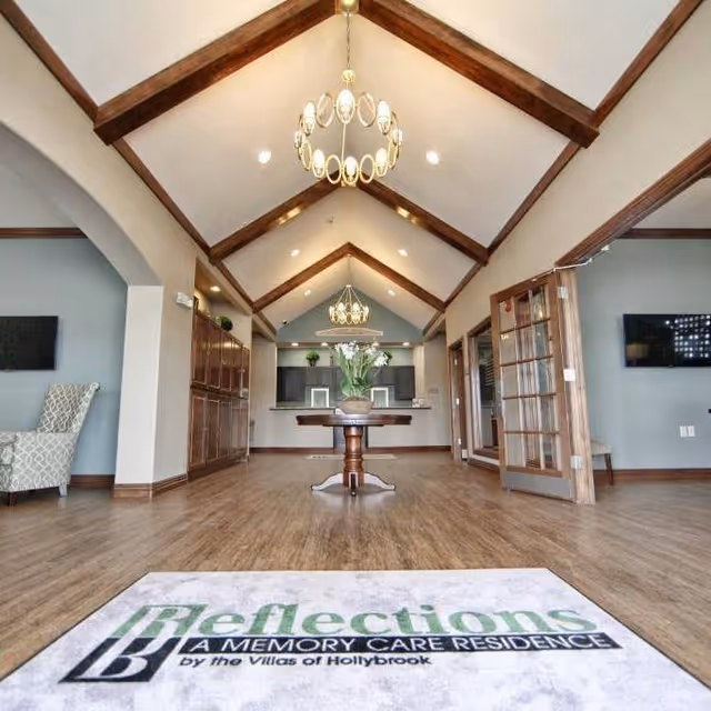 Spacious senior living lobby with vaulted beamed ceiling, chandeliers, a central table with flowers, seating, and an entry mat reading "Reflections A MEMORY CARE RESIDENCE by the Villas of Hollybrook".