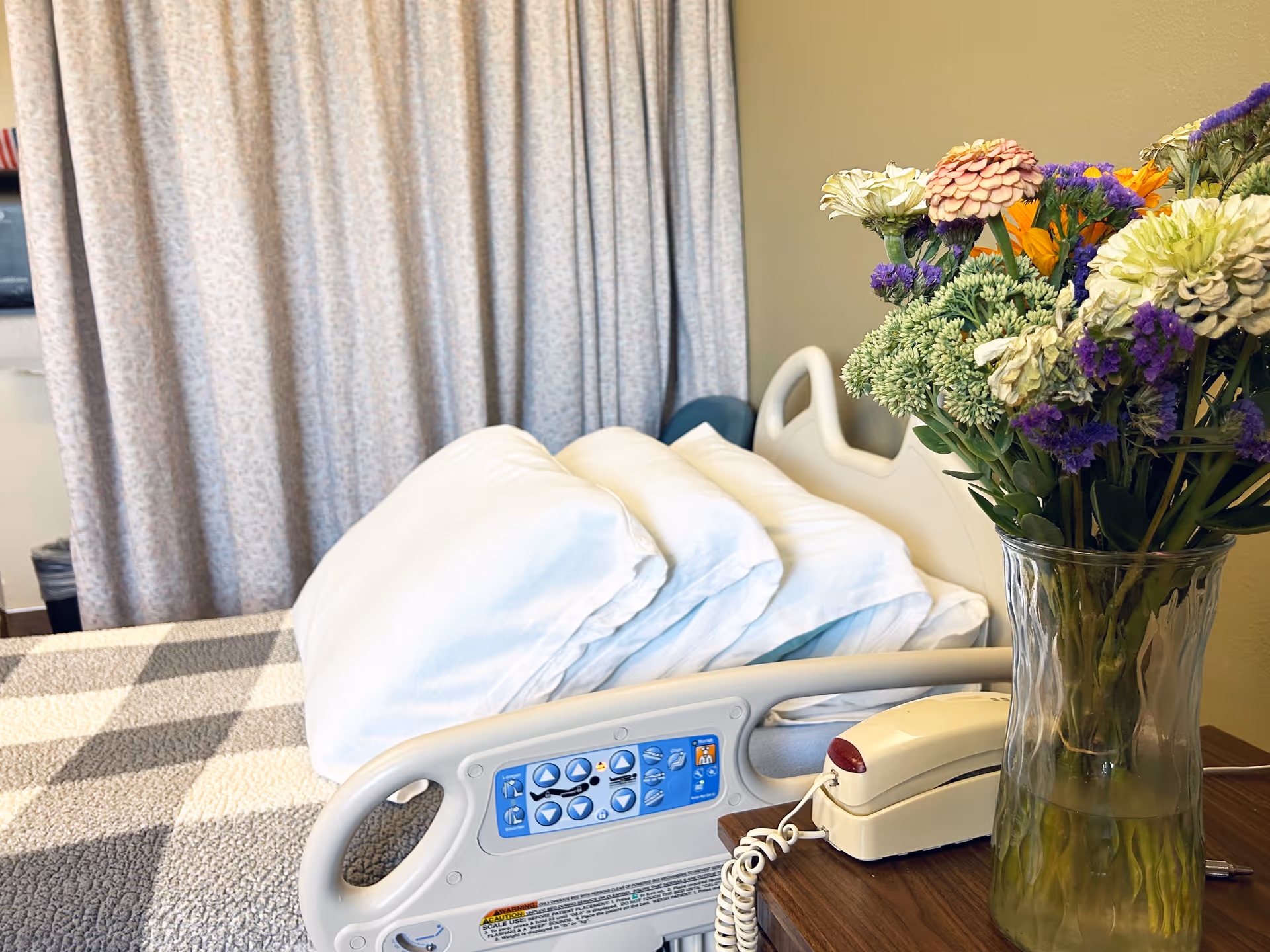 Hospital bed with flowers on a bedside table in a patient room.