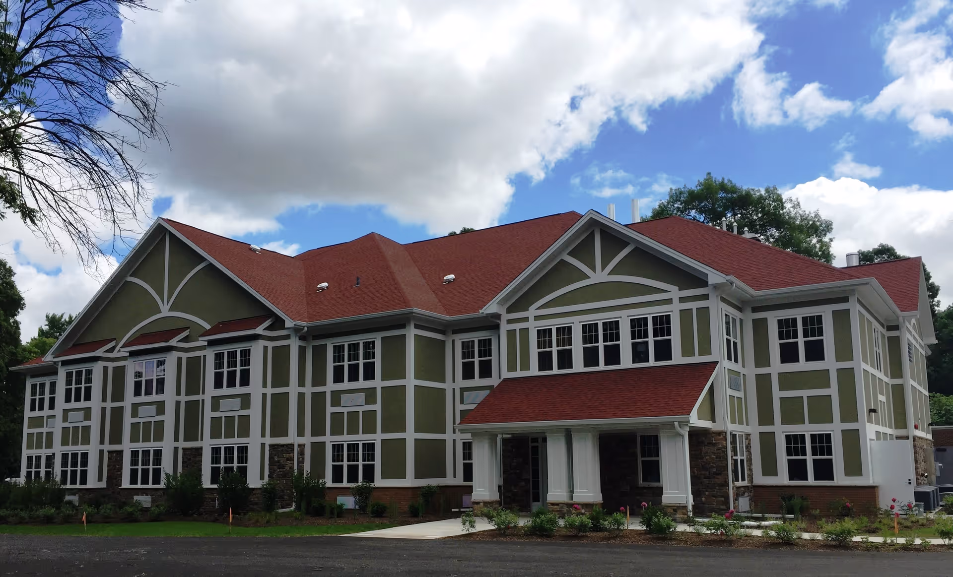 Exterior view of a large two-story senior living facility building with green and white walls and a red roof under a partly cloudy sky. The building has multiple windows and a covered entrance with columns, surrounded by some landscaping and trees.