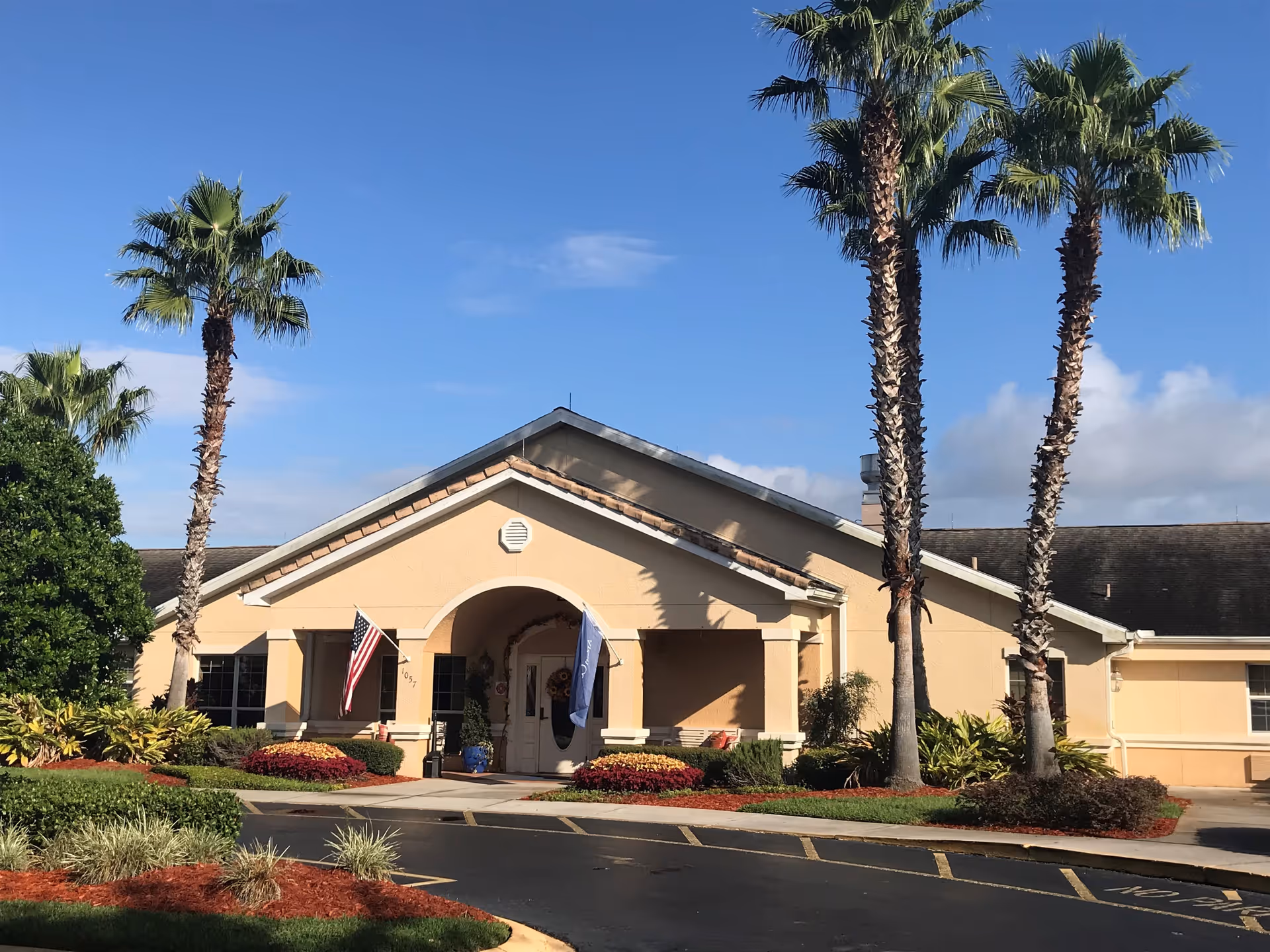 Exterior view of a single-story beige building with a peaked roof, palm trees, and landscaped bushes and flowers in front. Two flags, including an American flag, are displayed near the entrance under an archway. The sky is clear and blue.