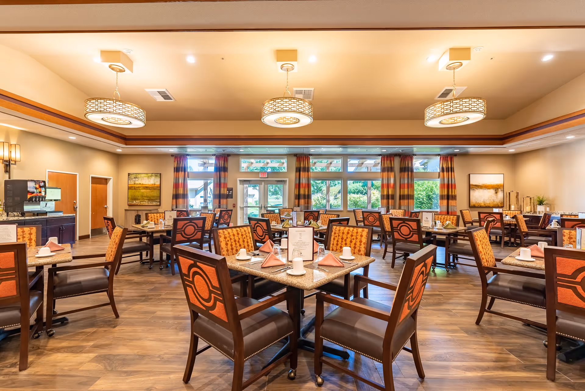 A bright and spacious dining room in a senior living facility with multiple tables set with cups, napkins, and menus. The room features wooden chairs with orange patterned cushions, large windows with striped curtains allowing natural light, and modern ceiling lights. The floor is wooden, and there are paintings on the walls.