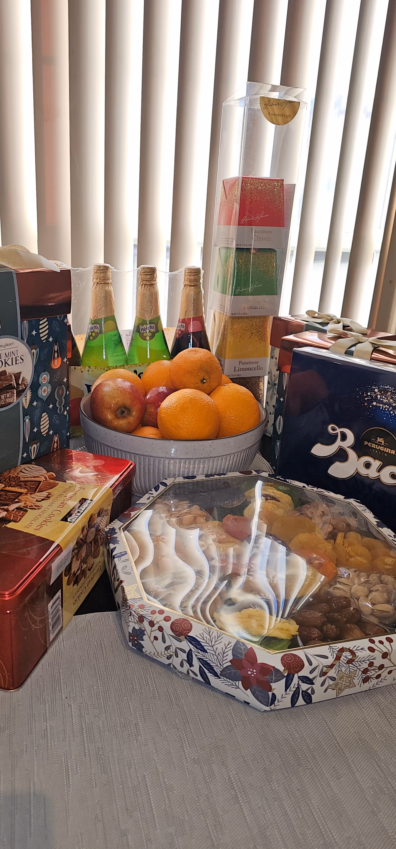 Table with assorted gifts, fruit, chocolates, and bottles in front of vertical blinds.