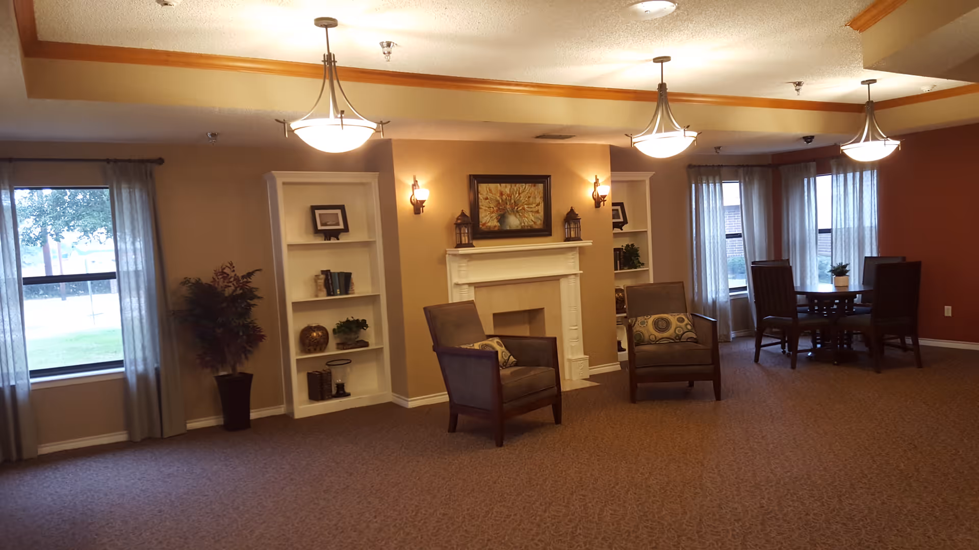 A cozy living room area with two armchairs facing a white fireplace. The fireplace mantel is decorated with two lanterns and a framed painting above it. Built-in white shelves flank the fireplace, holding books and decorative items. There are windows with sheer curtains on both sides of the room, and a round table with four chairs is visible near the windows on the right. The room has warm lighting from ceiling fixtures and wall sconces.