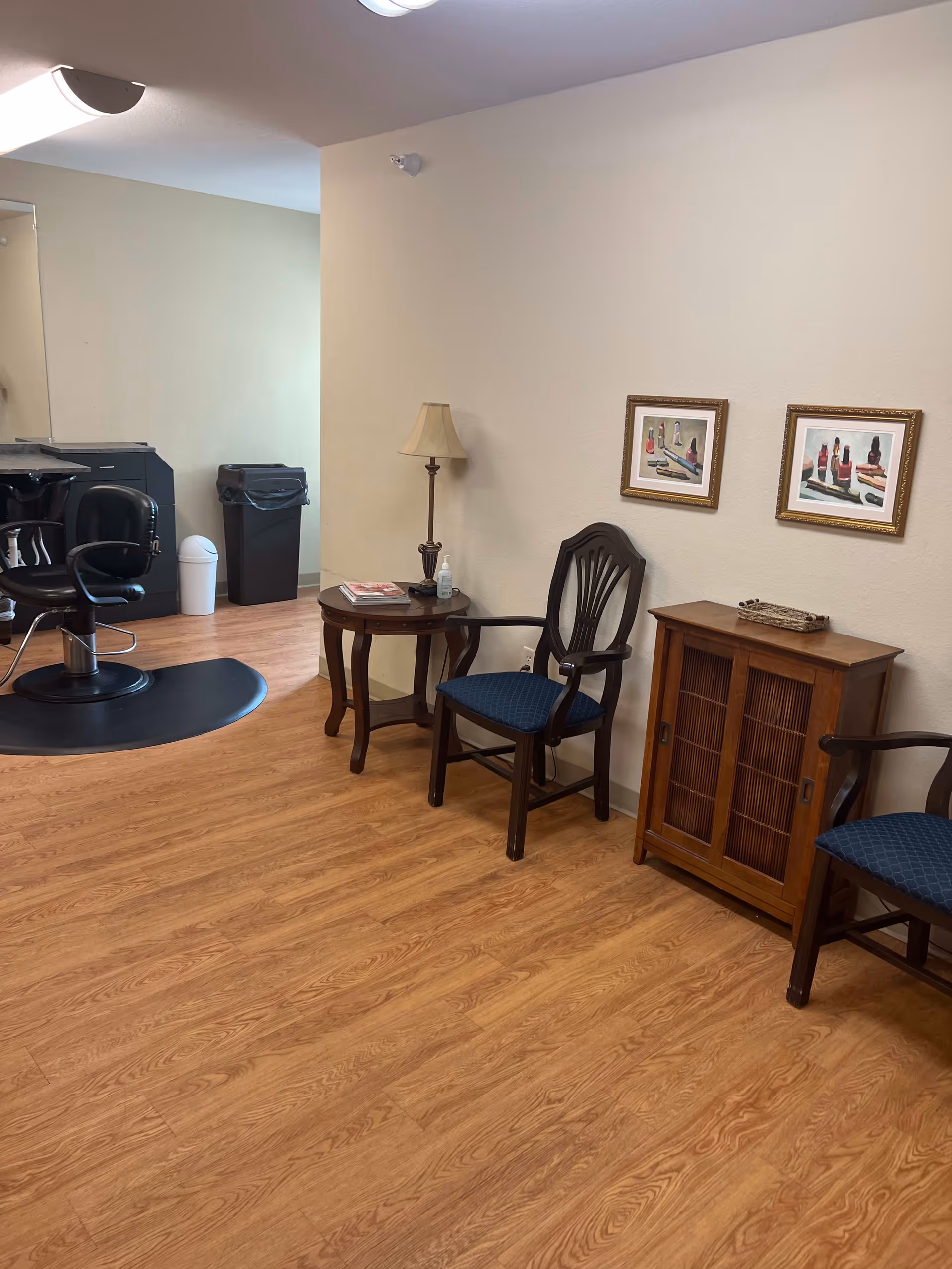 Interior view of a room with wooden flooring, two wooden chairs with blue cushions, a small wooden cabinet, a round wooden side table with a lamp and some items on it, two framed pictures on the wall, and a black salon chair with a black mat underneath. There are also two trash bins in the background.