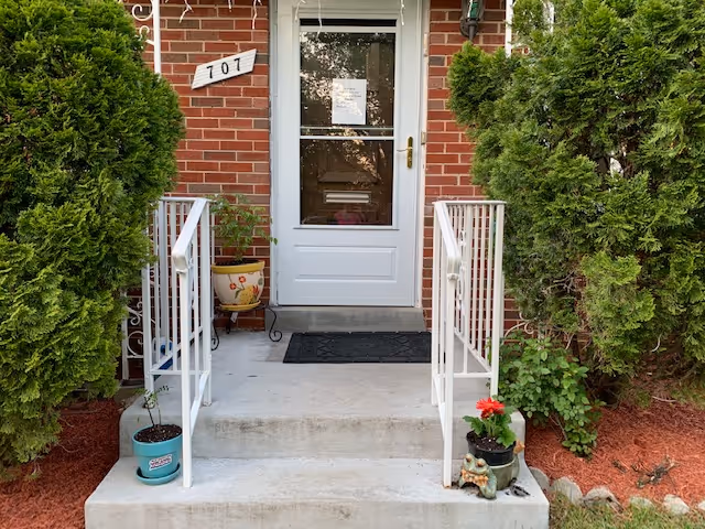 Front entrance of a brick building with a white door, steps, railings, and potted plants.