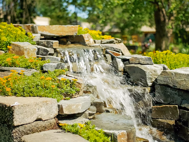 Stone outdoor waterfall feature cascades over rocks with surrounding landscaping.