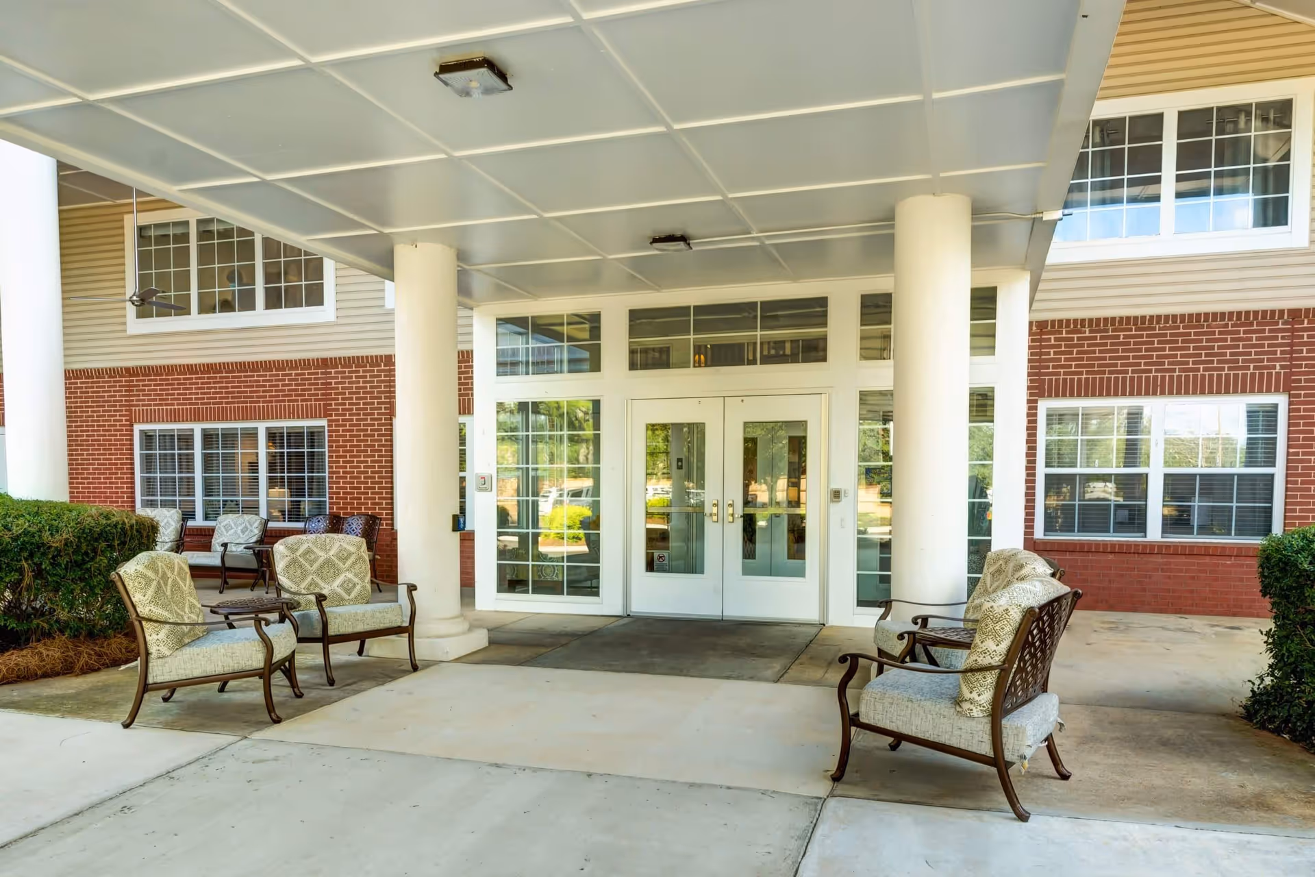 Covered entrance area of Ridgeland Place with white double doors, large white columns, and cushioned outdoor chairs arranged on either side of the walkway. The building exterior features red brick and beige siding with multiple windows.