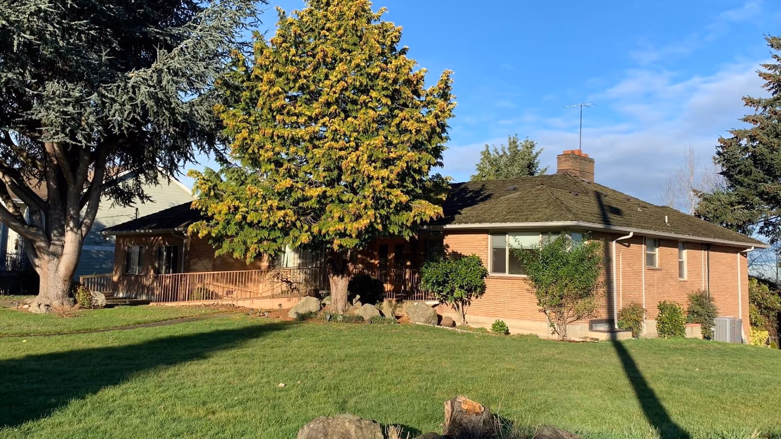 Single-story brick house with a green front lawn, trees, and a ramped porch under a blue sky.