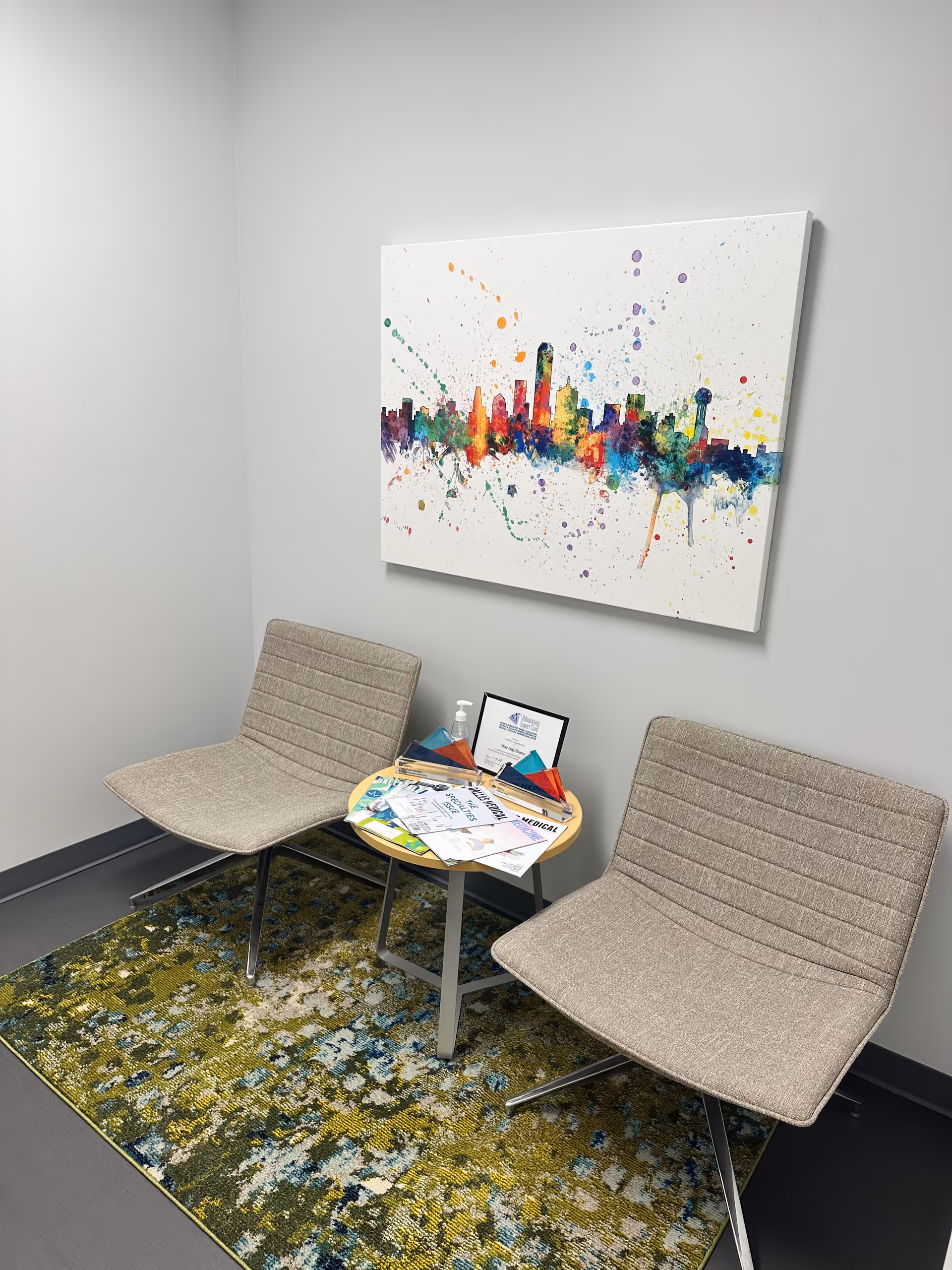 Two beige lounge chairs flank a small round table with magazines in a waiting area.