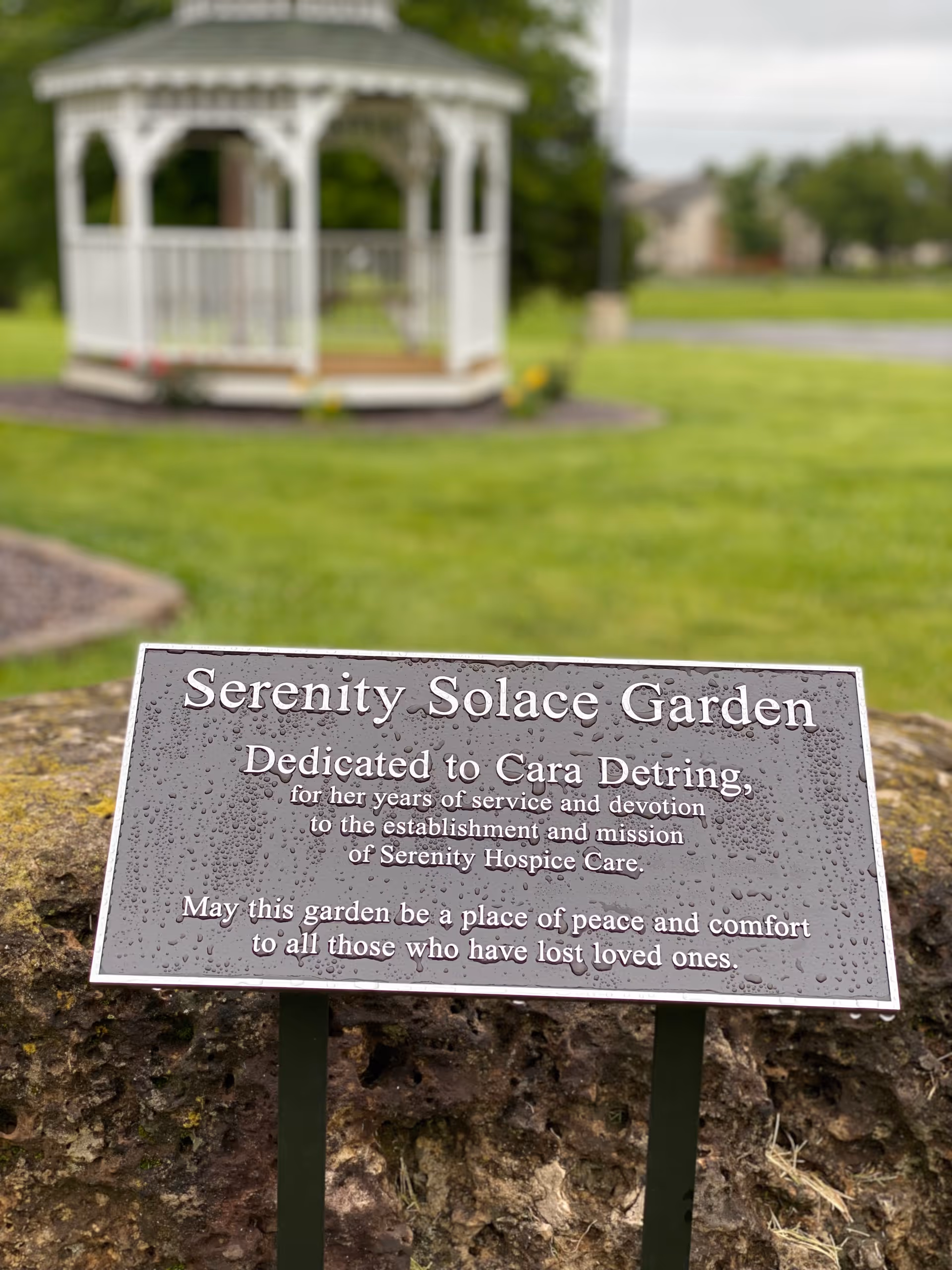 A white gazebo sits in a manicured lawn with a Serenity Solace Garden sign.