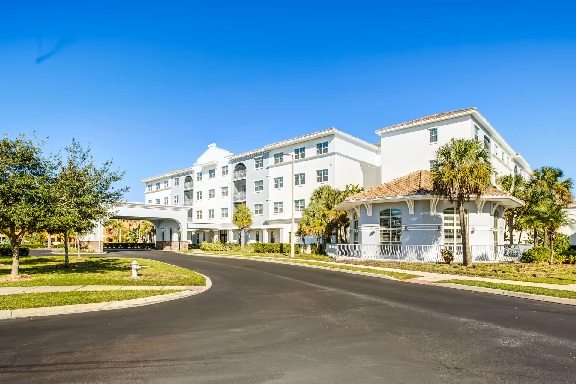 White multi-story senior living building with a curved driveway and palm trees at entrance
