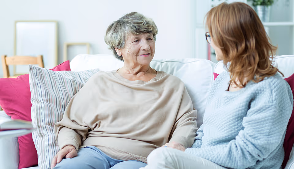 Two elderly women sit on a sofa in a bright living room, smiling and talking.