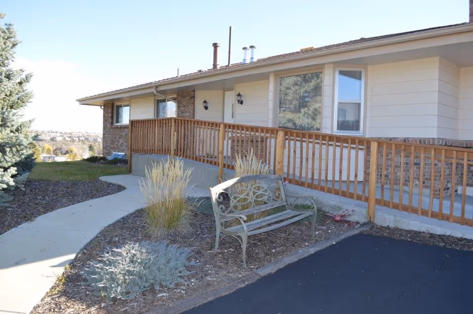 Front exterior of a single-story assisted living building with a wooden ramp, bench, and landscaped walkway.