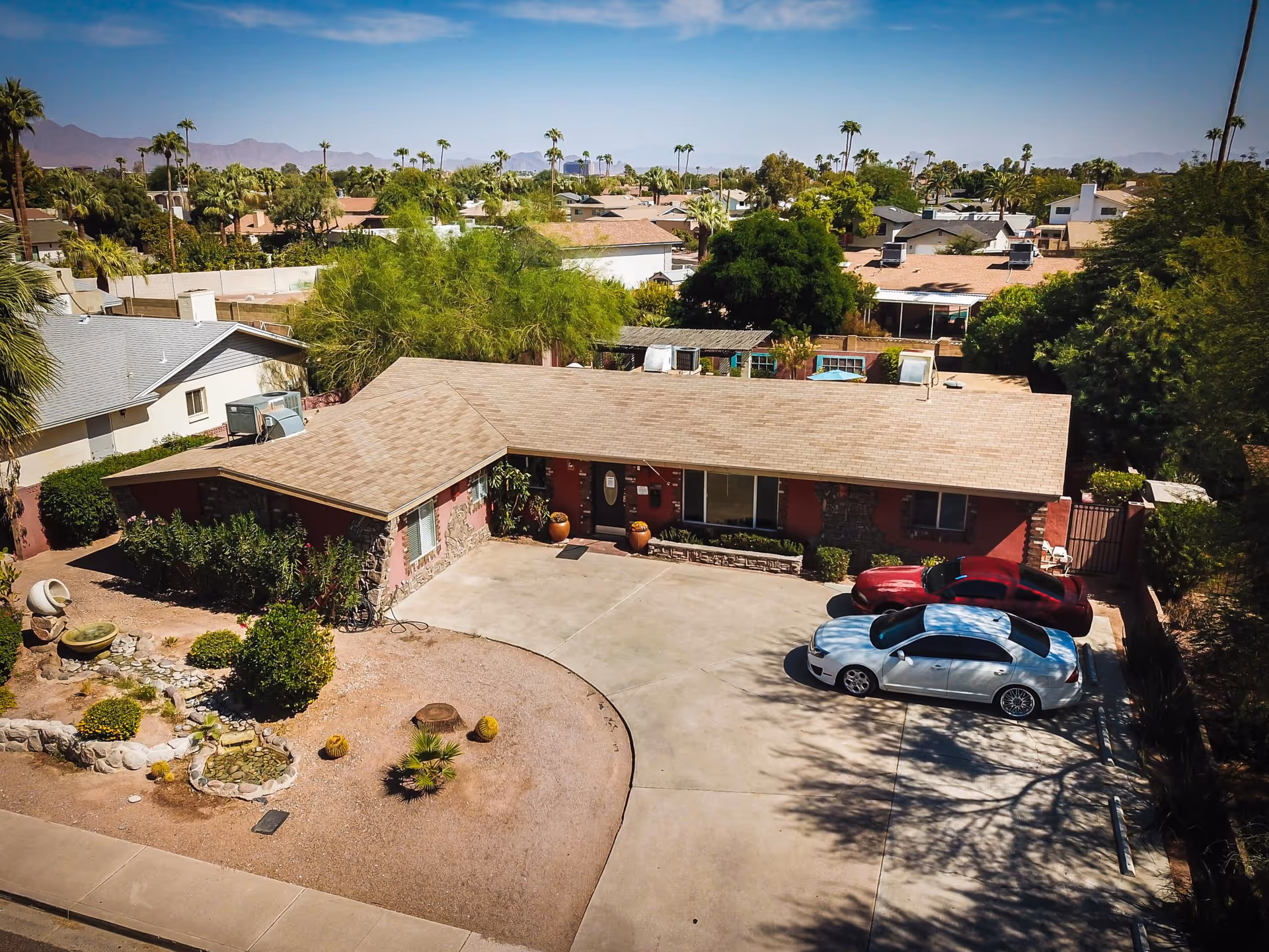 Aerial view of a single-story residential building with a beige roof and red exterior walls, surrounded by desert landscaping with small bushes and cacti. Two cars, one white and one red, are parked in the concrete driveway in front of the building. The background shows neighboring houses, palm trees, and distant mountains under a clear blue sky.