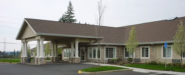 Exterior view of Cedar Crest Alzheimer's Special Care Center showing a single-story building with a covered entrance supported by stone and white columns, surrounded by a parking lot and small trees.